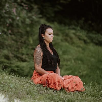 Donna con tatuaggio meditando su un prato verde, indossando un top nero e una gonna arancione.