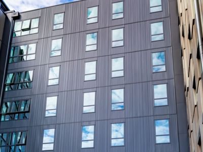 Modern multi-story building facade with vertical gray paneling and rectangular windows reflecting blue sky at Tupuārangi.