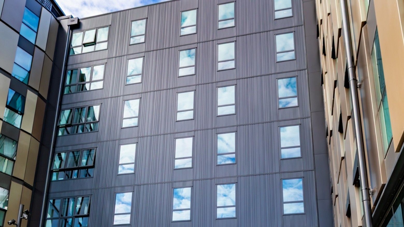 Modern multi-story building facade with vertical gray paneling and rectangular windows reflecting blue sky at Tupuārangi.