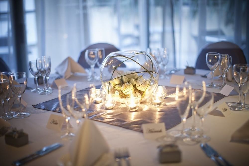 An elegantly set dinner table with a glowing floral centerpiece in a glass bowl and lit candles at The Sebel Brisbane