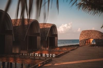 Seaside view of arch-roofed villas at Ananea Madivaru Maldives, with wooden walkways over calm waters and a pastel sky