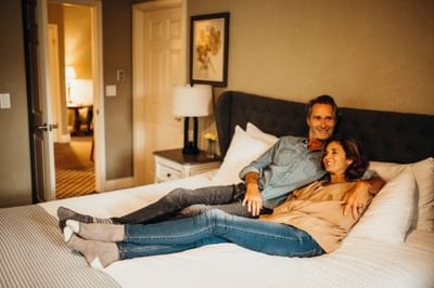 Couple lounging on a bed with a nightstand and lamp beside an open door in a hotel room.