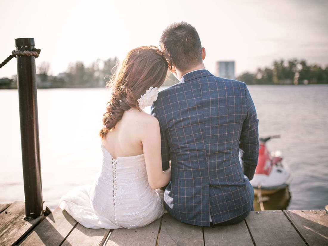 Couple in wedding attire sits on a dock overlooking a lake, capturing a beautiful moment at The Sebel Mandurah