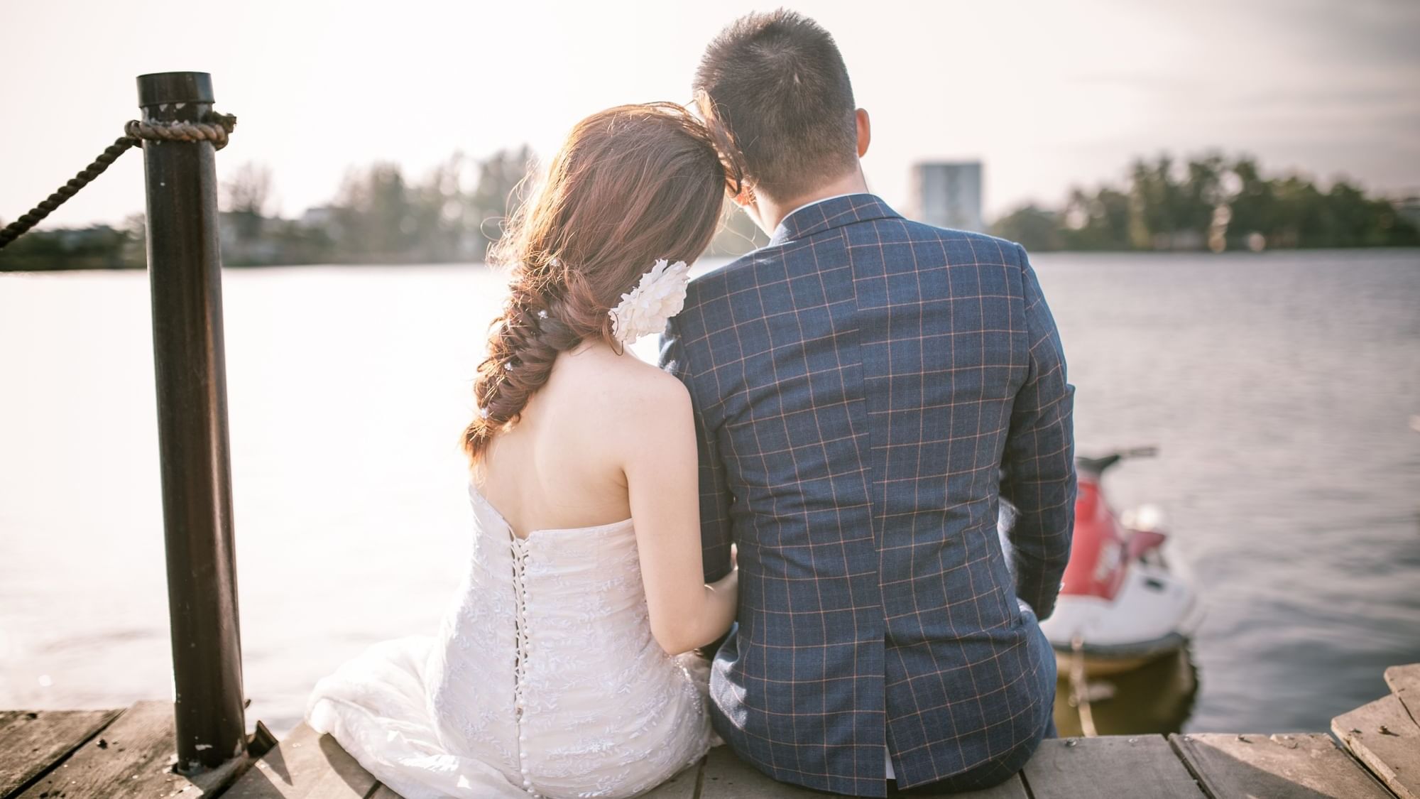 Couple in wedding attire sits on a dock overlooking a lake, capturing a beautiful moment at The Sebel Mandurah