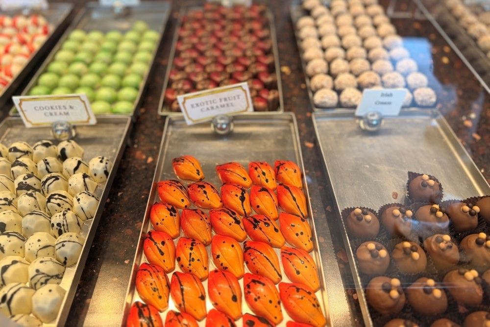 A close-up of a display case of colorful handmade chocolates, including orange exotic fruit caramels splattered with black and an assortment of others.