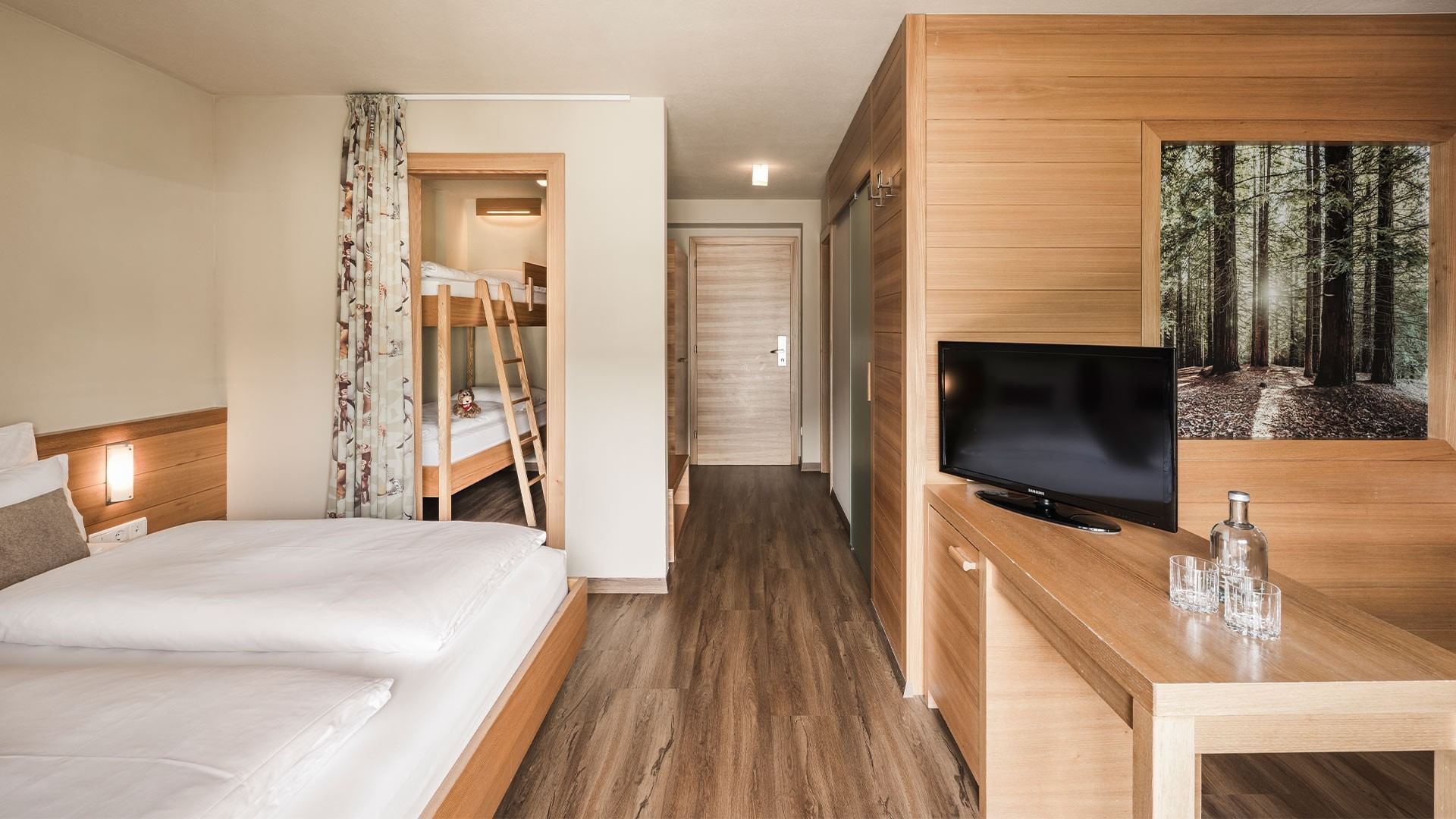 Hotel room with wooden floors, two beds, a desk with a TV, and a bunk bed in the background.