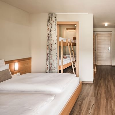 Hotel room with wooden floors, two beds, a desk with a TV, and a bunk bed in the background.