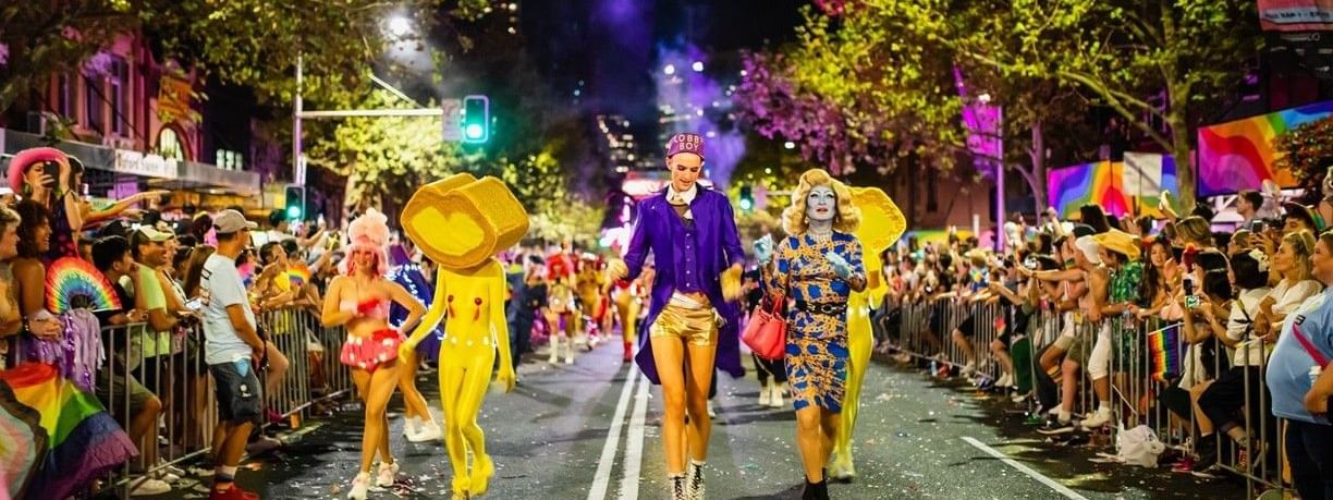 Performers in vibrant costumes walk down a city street lined with spectators at the Sydney Mardi Gras 2026.