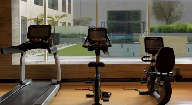 Modern gym with a treadmill and stationary bikes facing glass windows overlooking the lawn at DAMAC Maison Cour Jardin