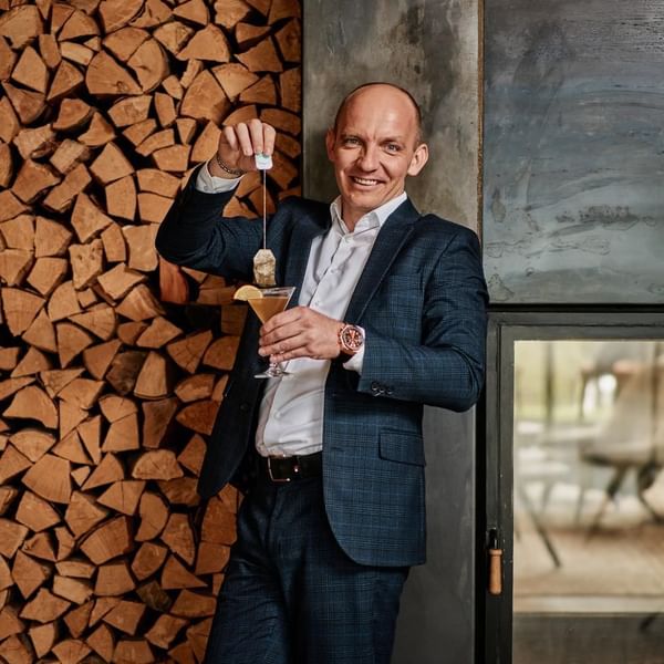 Mann in Anzug mit Cocktailglas, der Teebeutel hineinhängt, vor einem Hintergrund aus Holz und Metall.