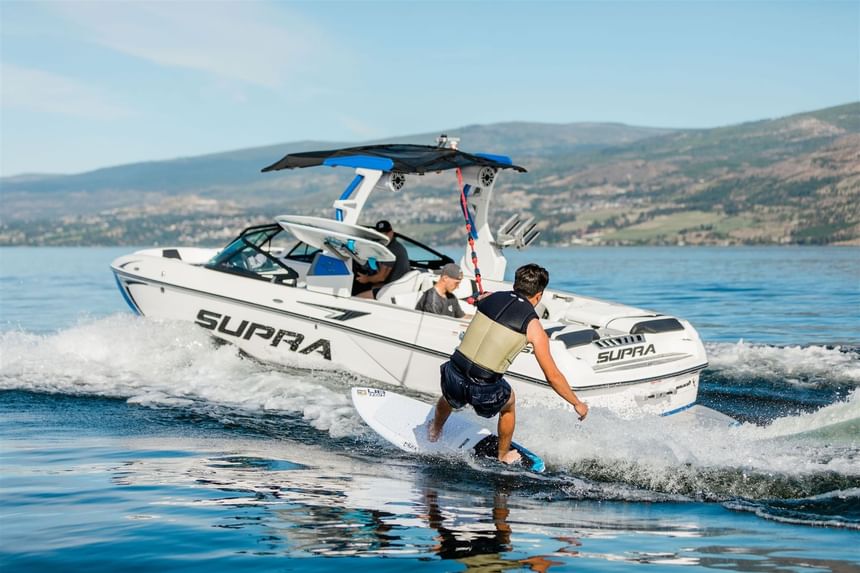 A man surfs behind a SUPRA boat on the water with mountains in the background.