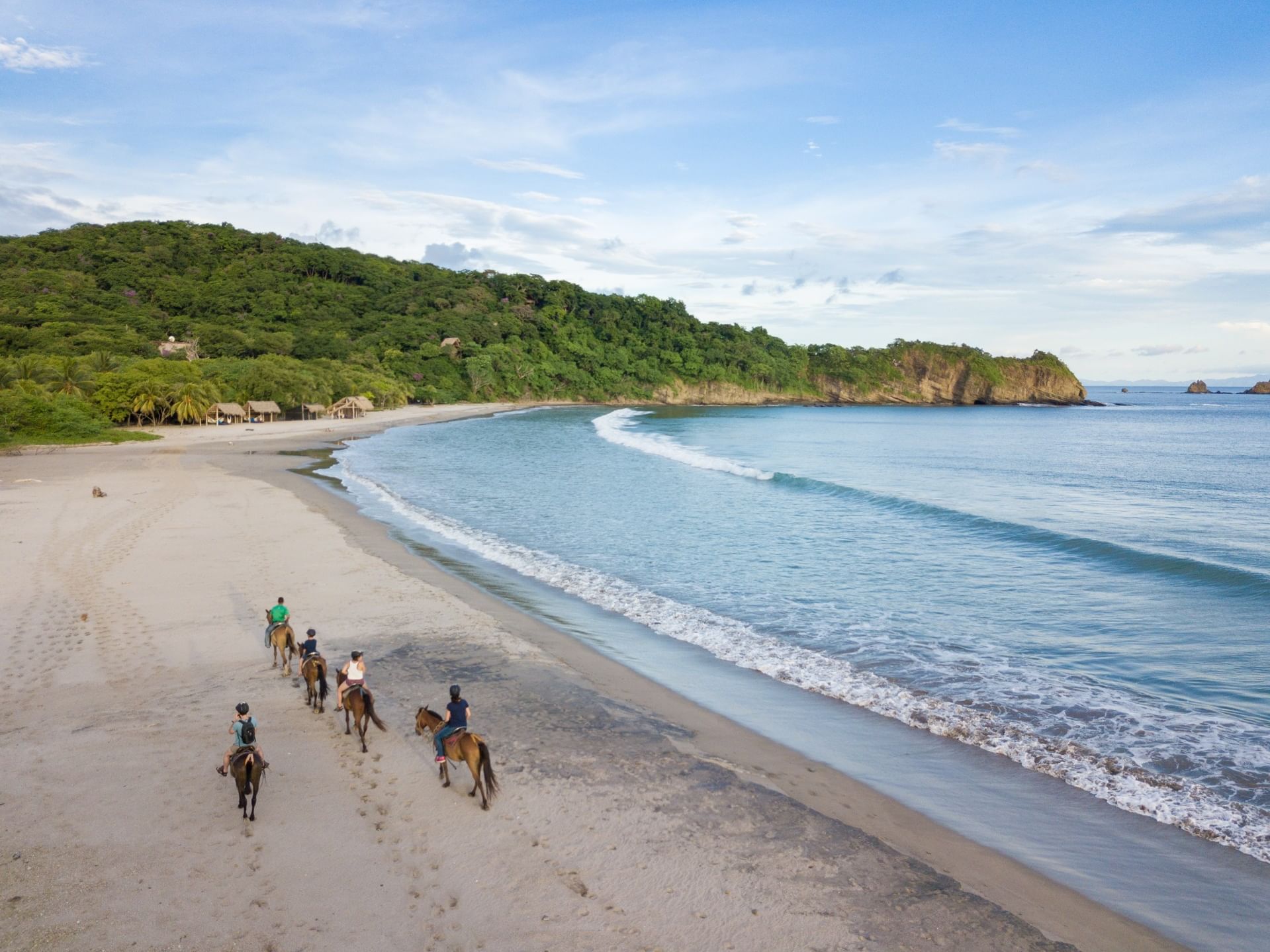 Group of riders on horses by the ocean waves under a blue sky on the beach at Morgan's Rock Reserve & Ecolodge