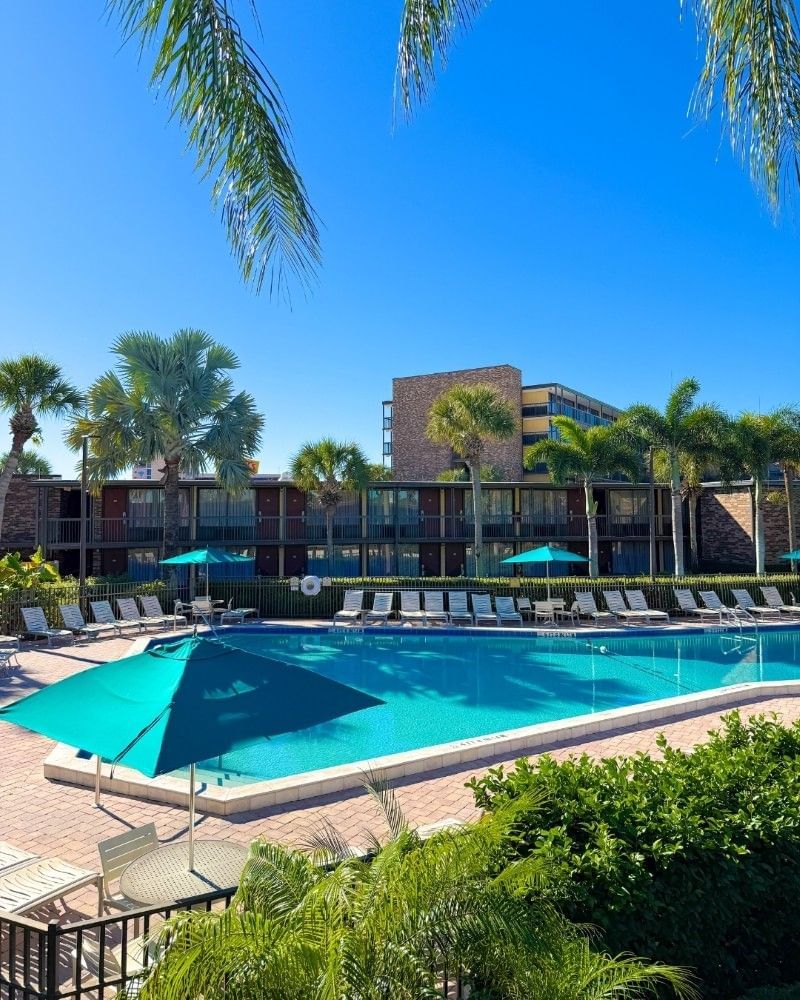 An angular pool on a sunny day surrounded by white lounge chairs, blue umbrellas, and a two-story hotel building at Rosen Inn International.