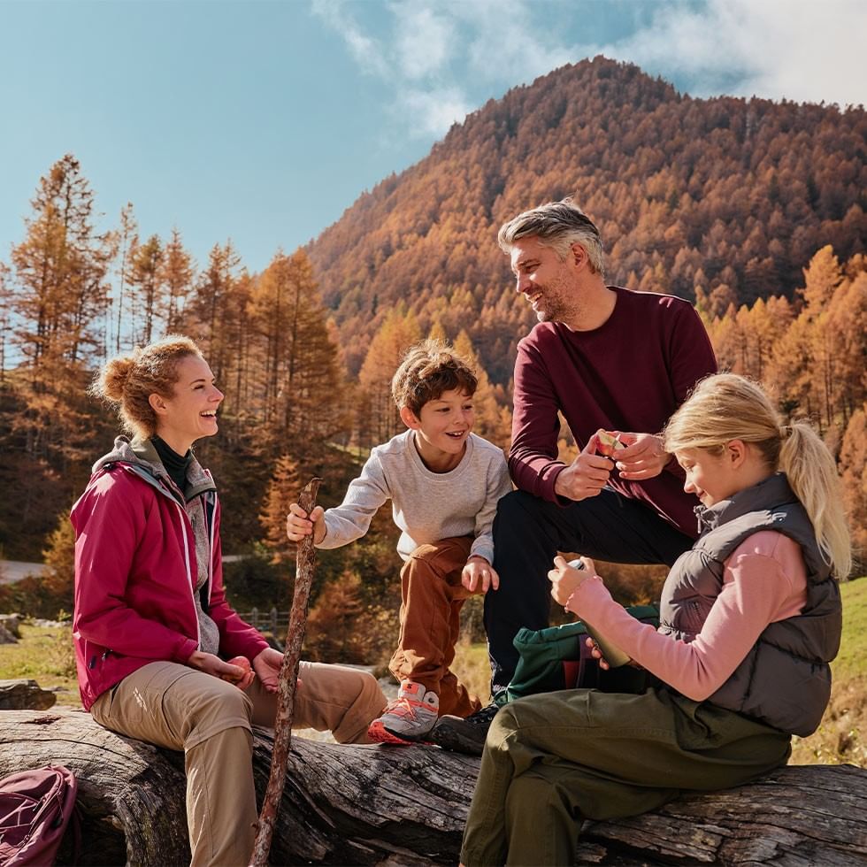 Vier Personen sitzen auf einem Baumstamm, umgeben von herbstlichen Bäumen und einem Berg.