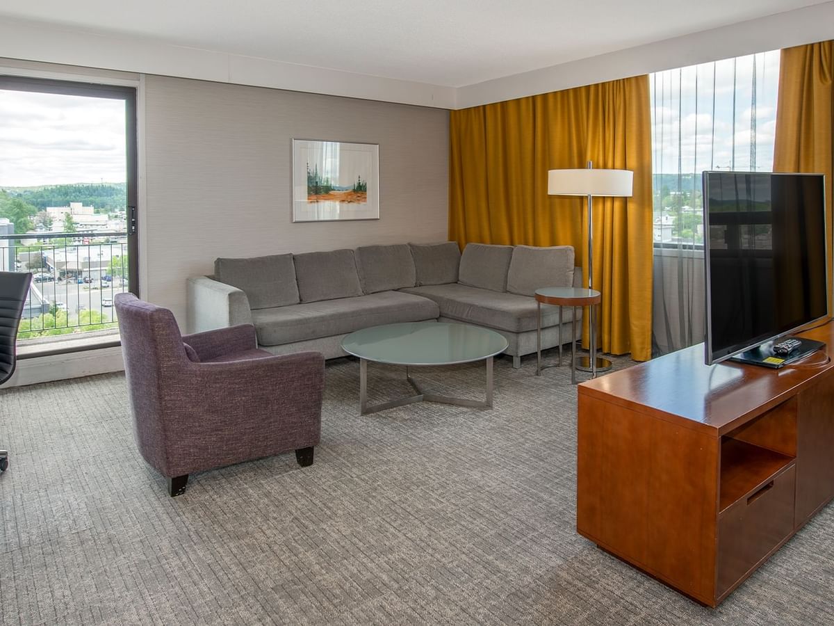A hotel suite living area with a couch, TV , purple chair, glass table, and a balcony view.