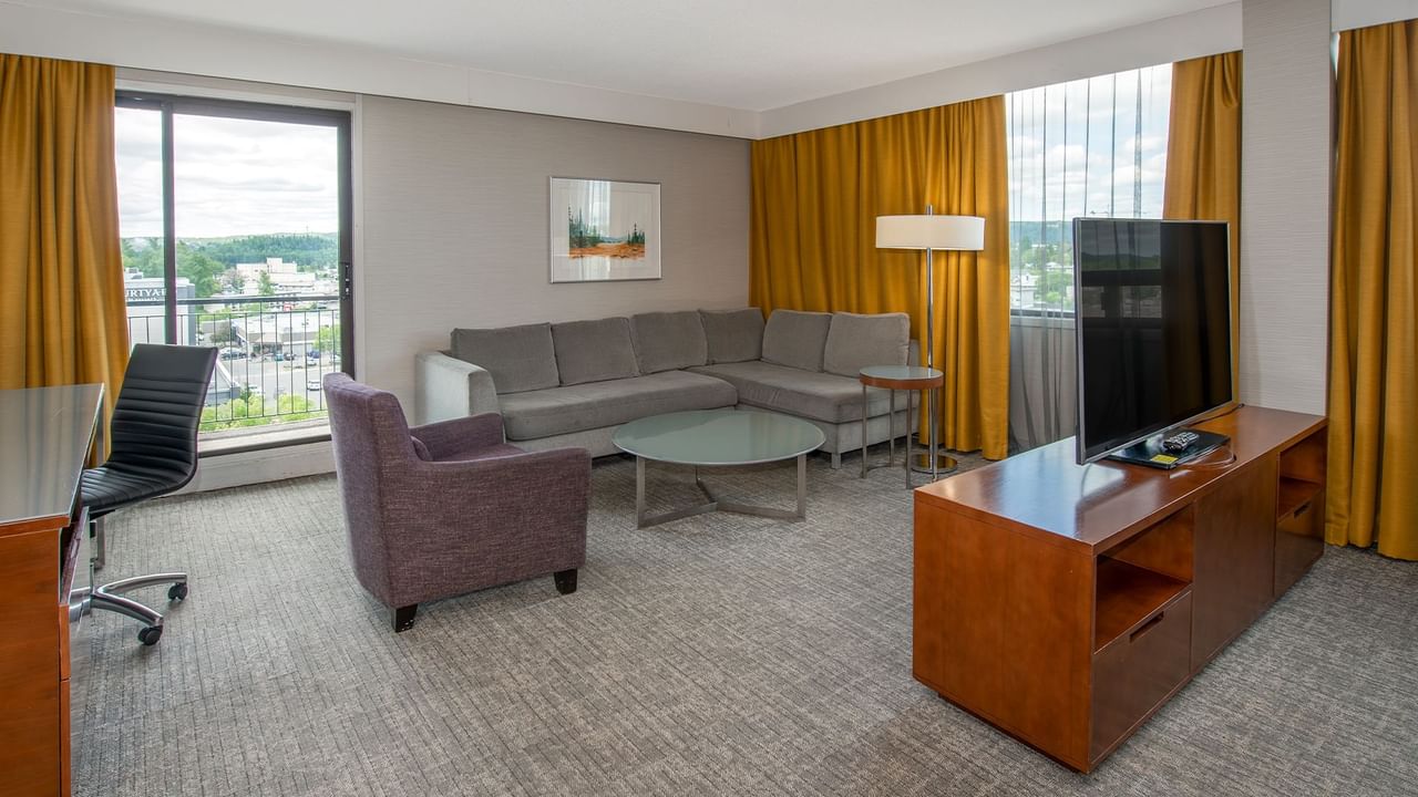A hotel suite living area with a couch, TV , purple chair, glass table, and a balcony view.