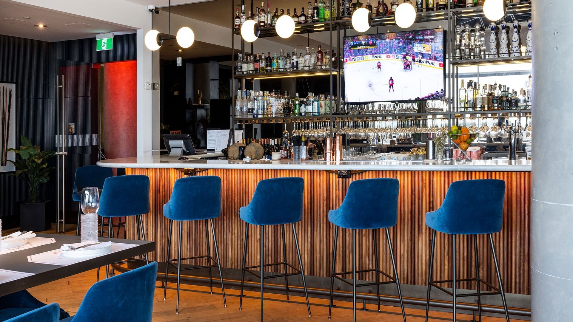 Roses Bar counter with blue velvet stools facing a lit display shelf and wall mounted TV at Hotel X Toronto
