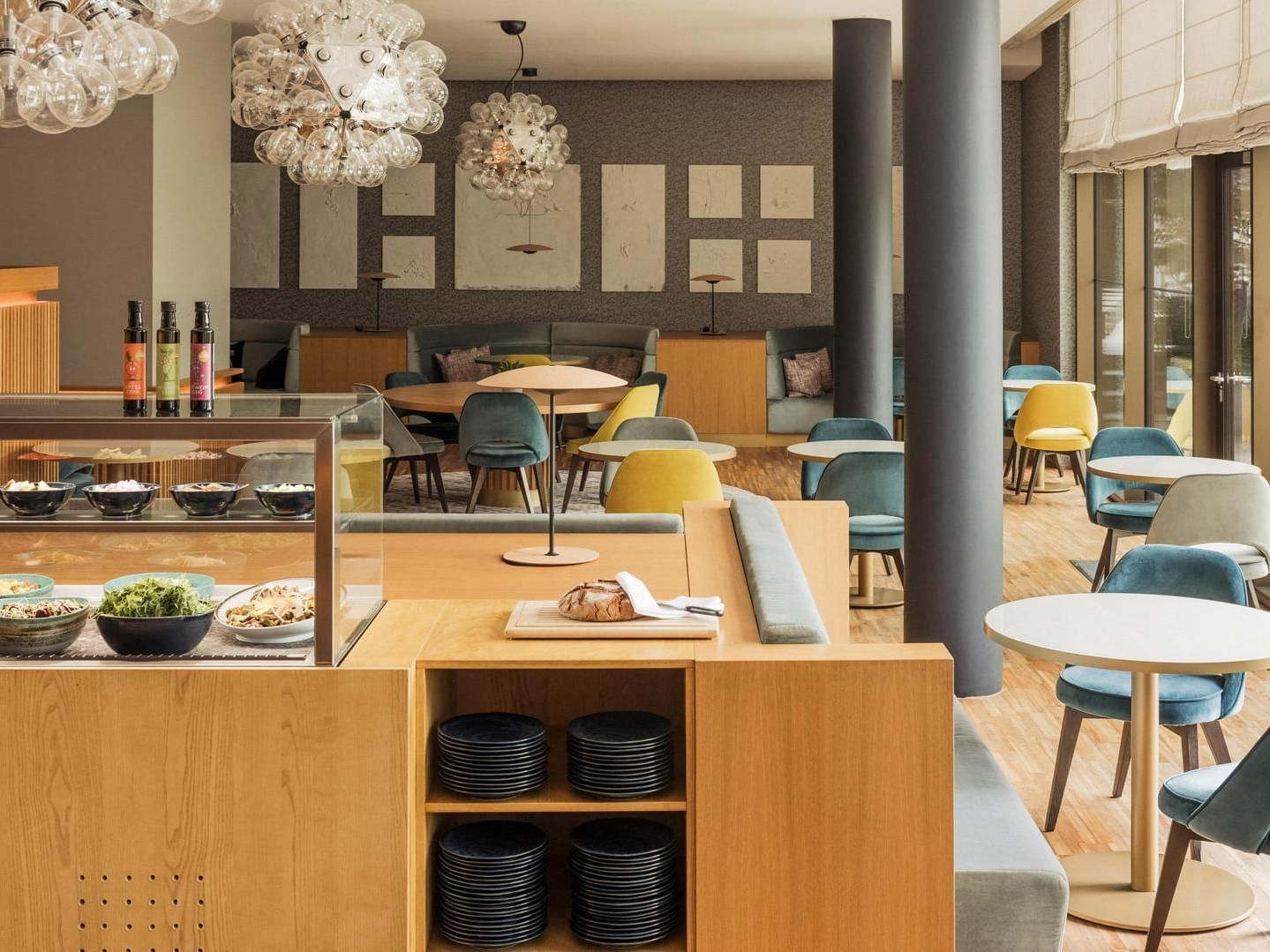 Modern cafe with wooden tables, colorful chairs, glass display with food, and decorative glass chandeliers.