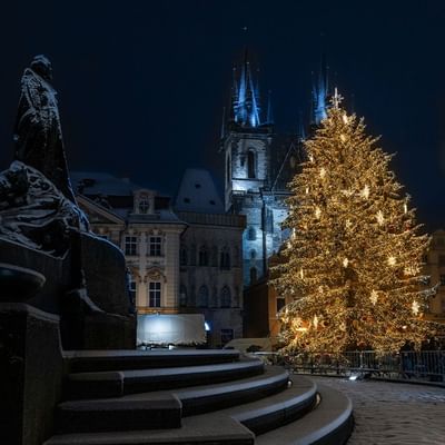Nachtszene mit beleuchtetem Weihnachtsbaum vor einer Kirche und einer Statue in einer schneebedeckten Stadt.
