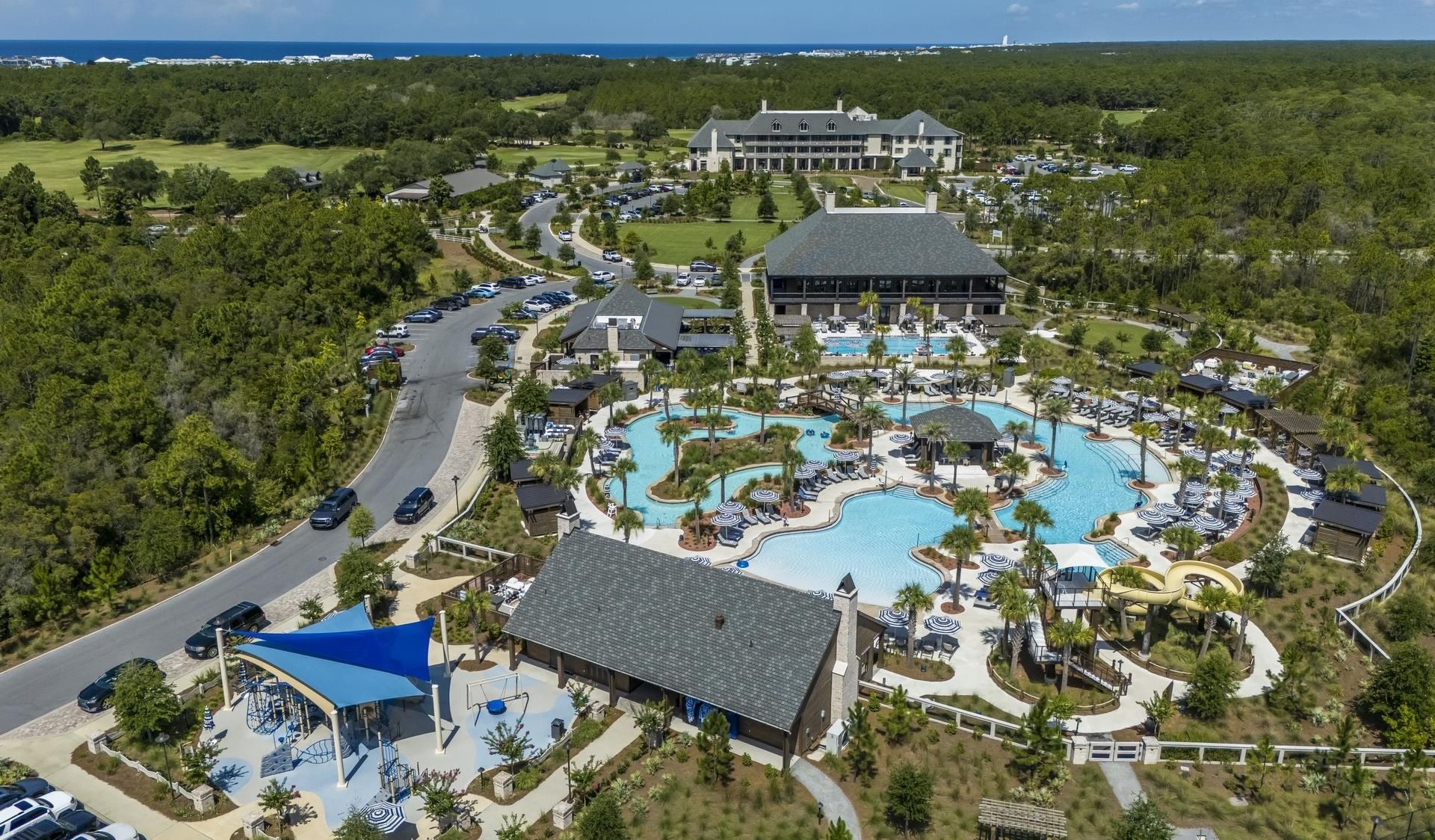 Aerial view of resort with buildings, pools, water slide, parking lot, and lush greenery.