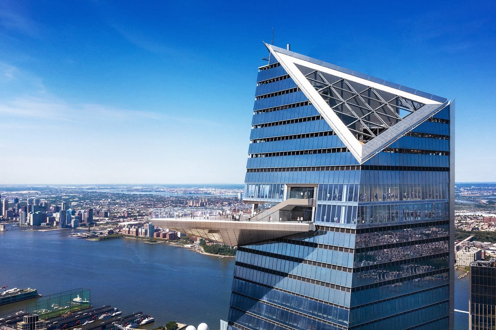 A blue glass building with a triangular roof overlooking a body of water and cityscape.