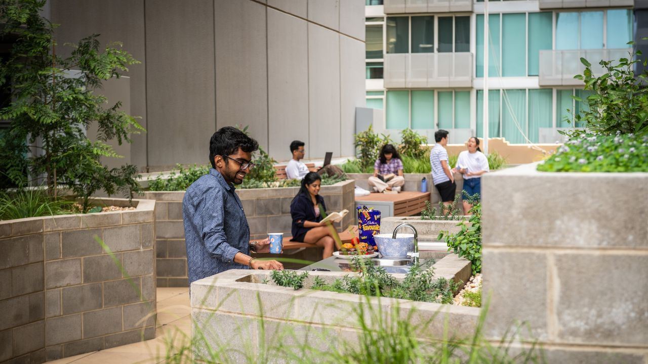 Man with glasses holds a cup in a outdoor terrace with raised garden beds and people.