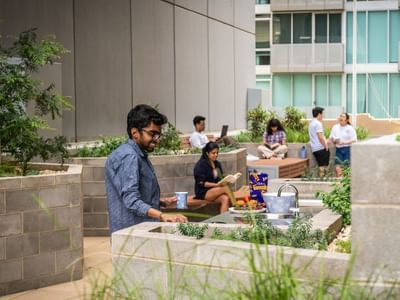 Man with glasses holds a cup in a outdoor terrace with raised garden beds and people.