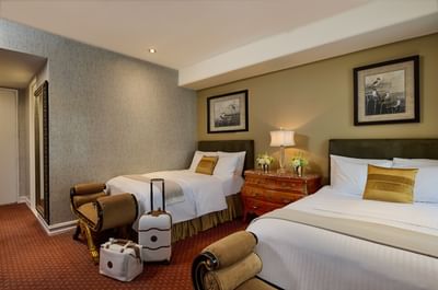 Two beds between wooden nightstand with lamp and white luggage on floor in double bedroom at The Kimberly Hotel