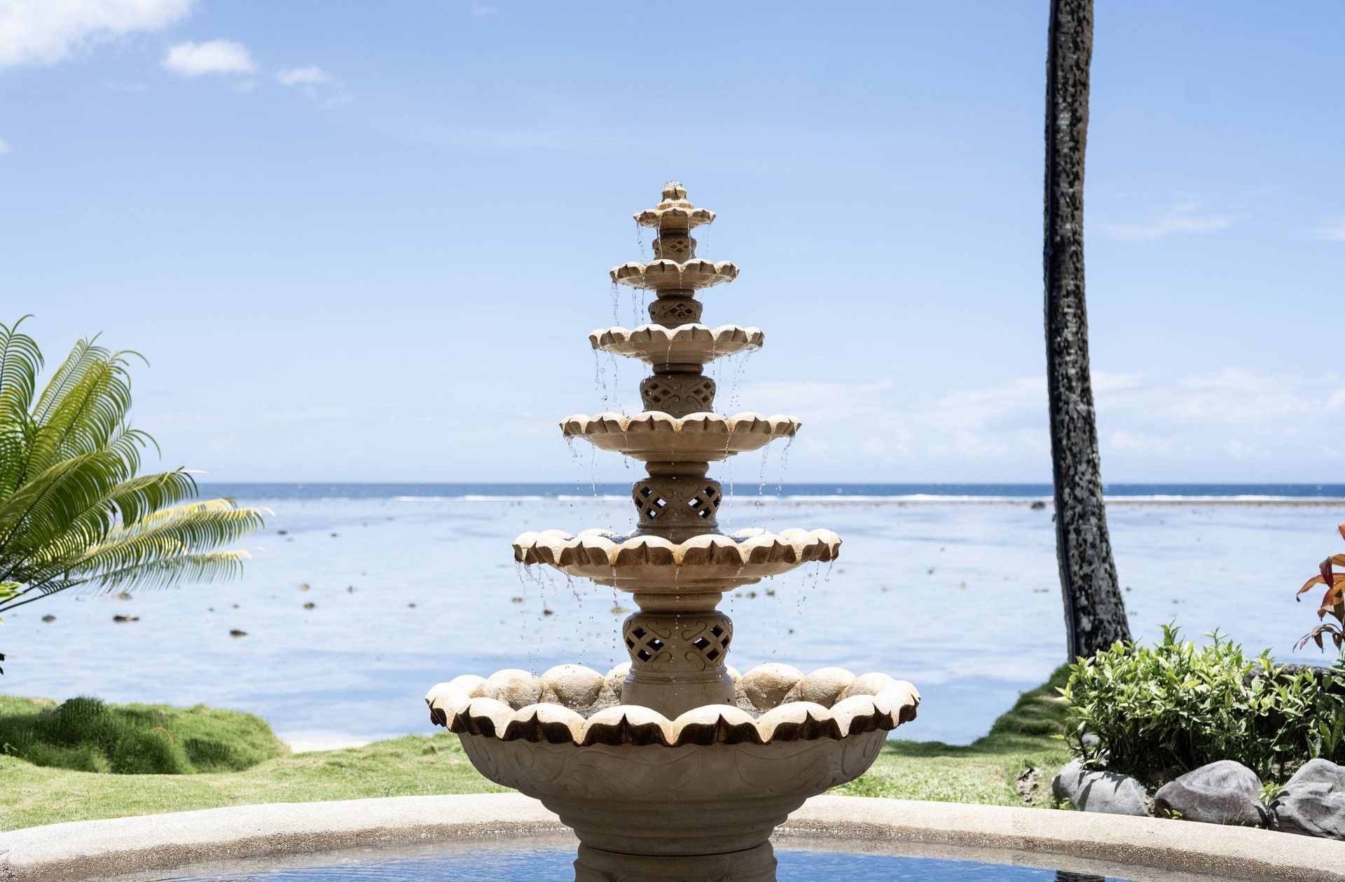 Tiered stone fountain by a palm tree under a bright sky surrounding a lagoon at Warwick Fiji Resort and Spa
