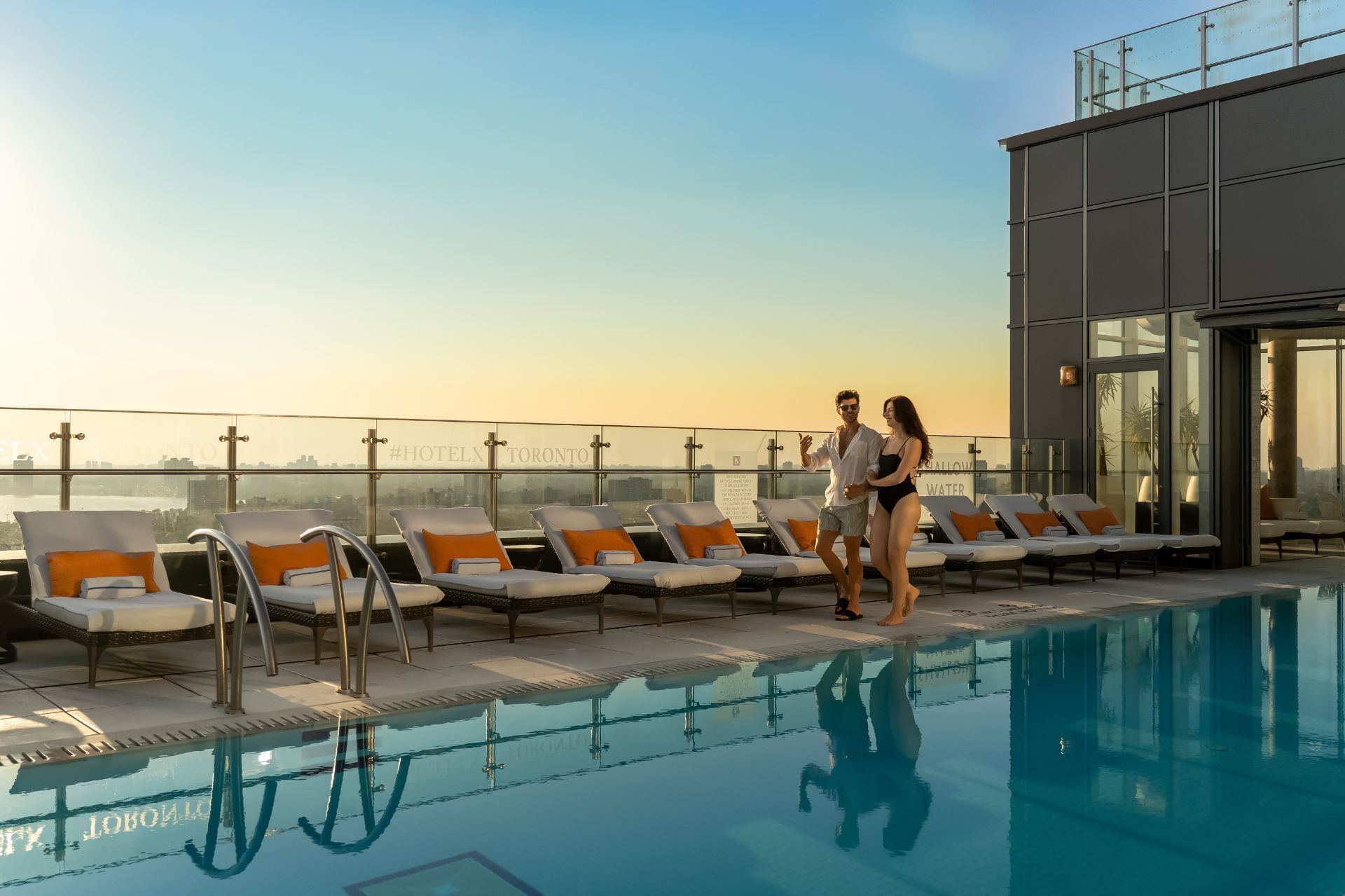 Couple walks by pool with lounge chairs on deck, overlooking cityscape at sunset.