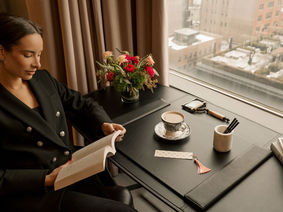 Woman reading a book at work desk with coffee cup and flowers by windows in Duplex Penthouse at Gansevoort Meatpacking NYC