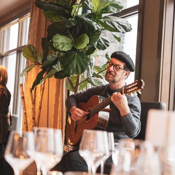 Man playing guitar near large plant and wine glasses in a room with large windows.