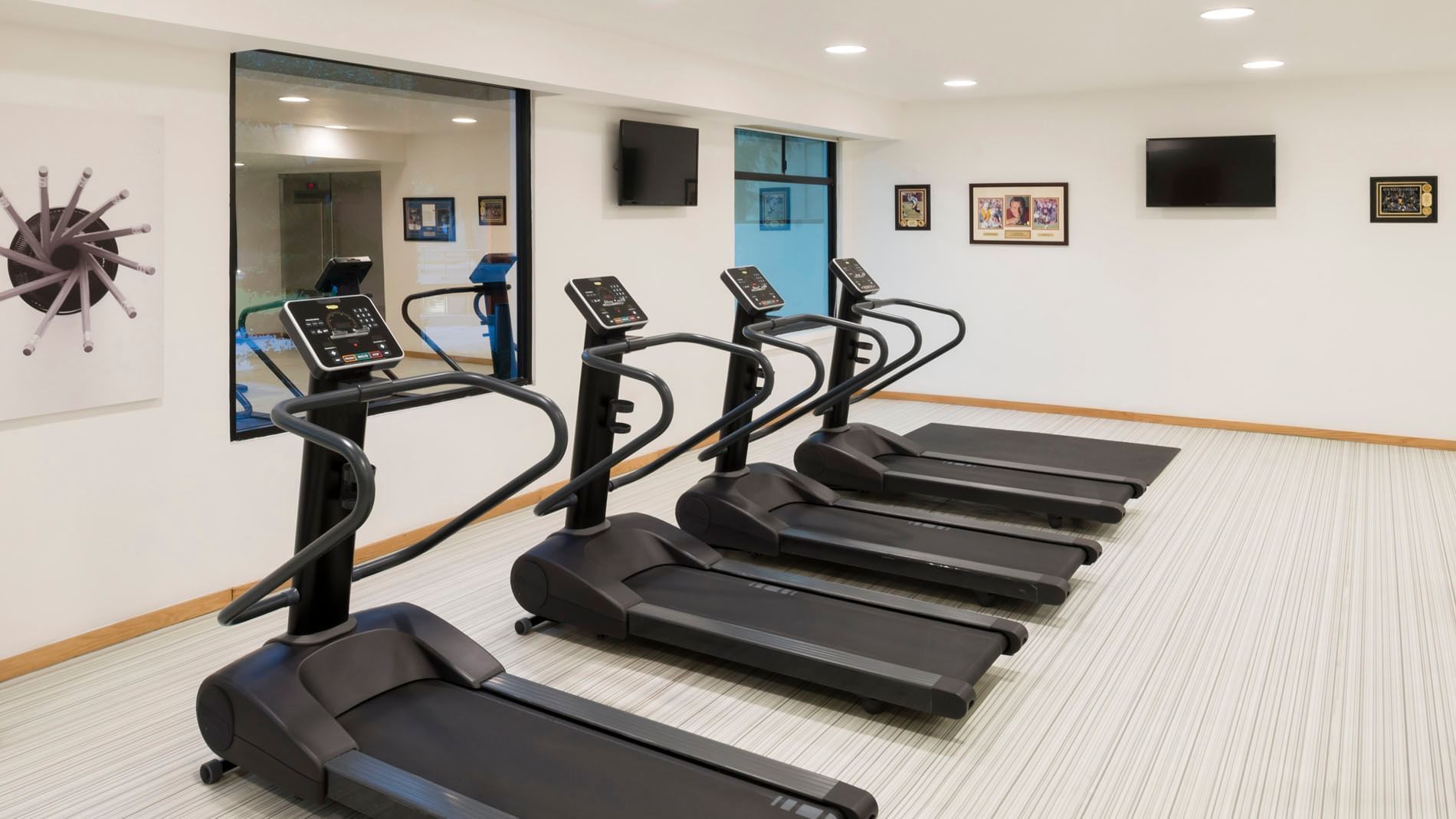 Black treadmills by a white wall under flat-screen TVs near a large window in the gym at the Real Inn Mexicali