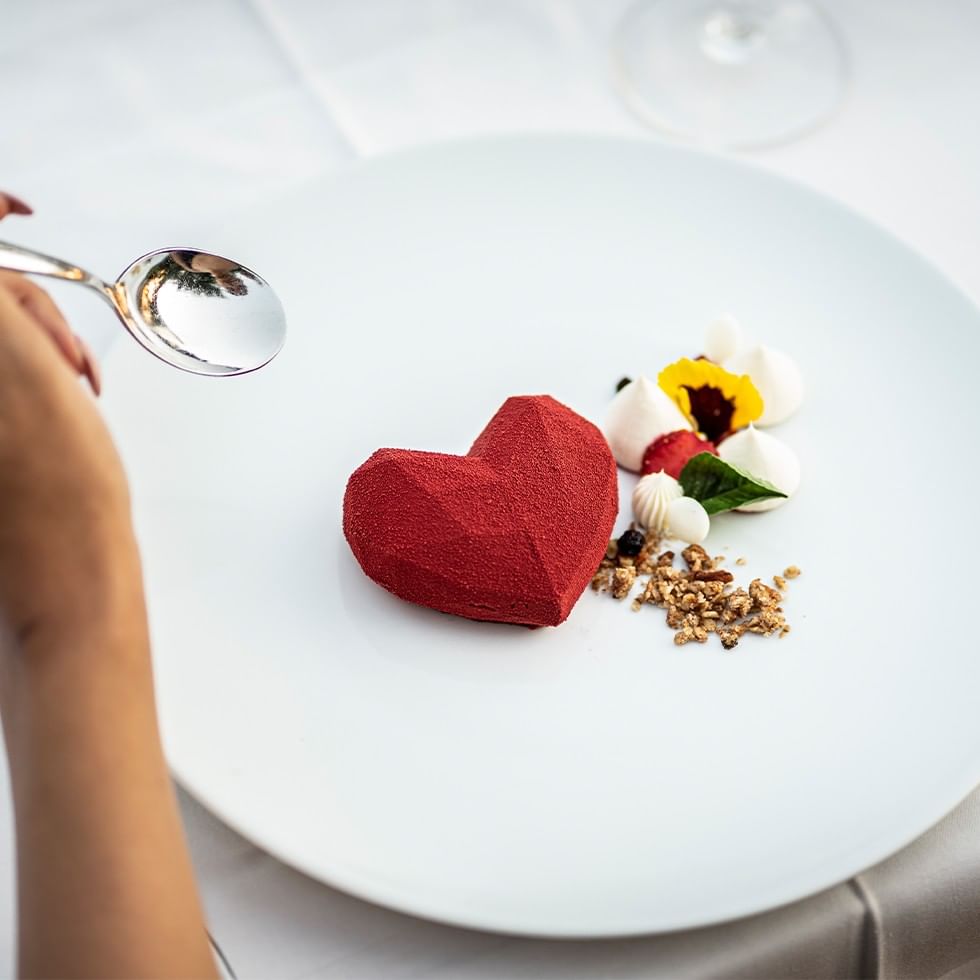 Hand holding a spoon next to a plate with a red heart-shaped dessert.