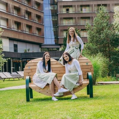 Drei Frauen in Dirndln sitzen und stehen auf einer Herz-förmigen Bank vor einem Gebäude.