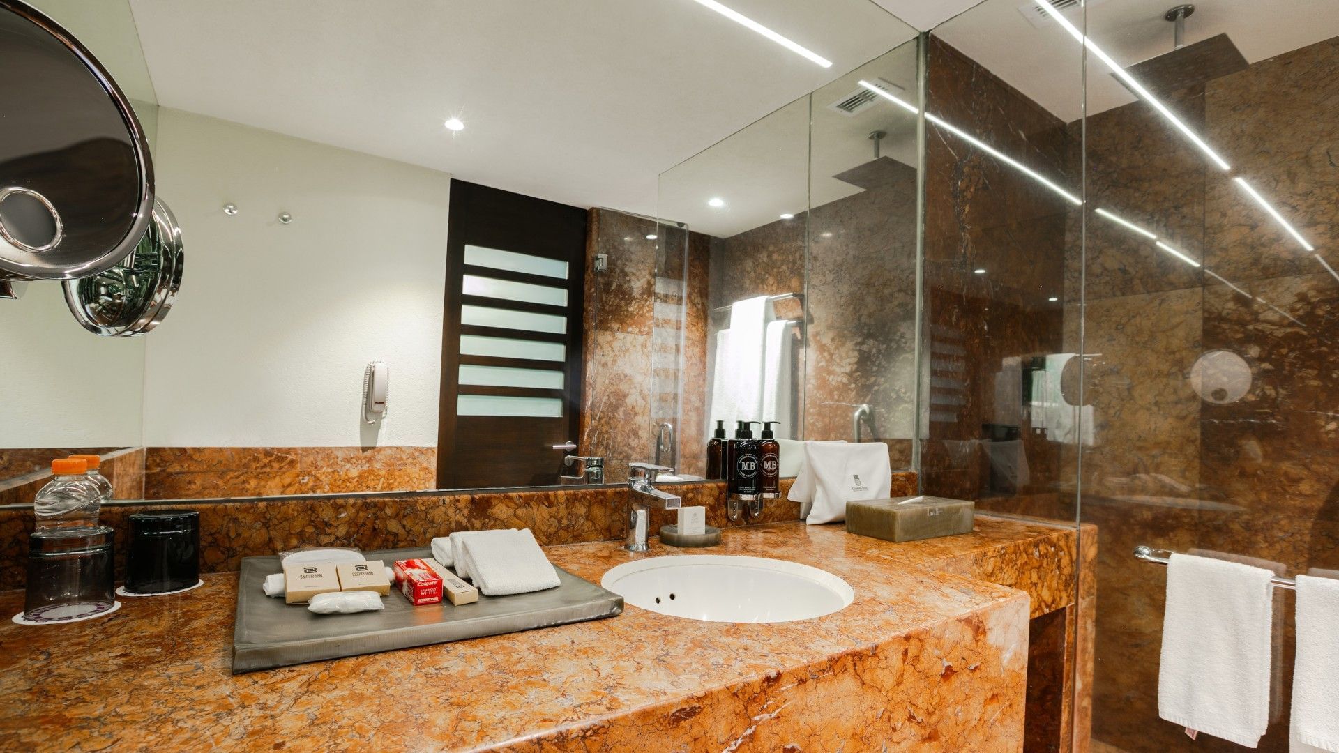 Baño elegante con espejo, lavabo, toallas, y ducha con puertas de vidrio y azulejos decorativos.