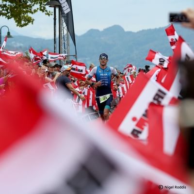 Athlet läuft durch jubelnde Menge mit Ironman-Fahnen in einer bergigen Umgebung.