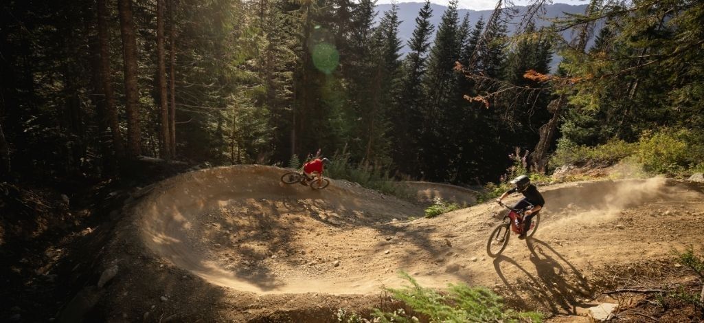 Two mountain bikers ride through a sunlit berm on a forest trail in Whistler.