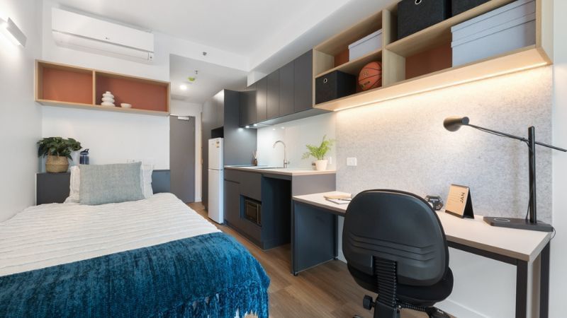 A studio apartment with a bed, desk, chair, and compact kitchen area.