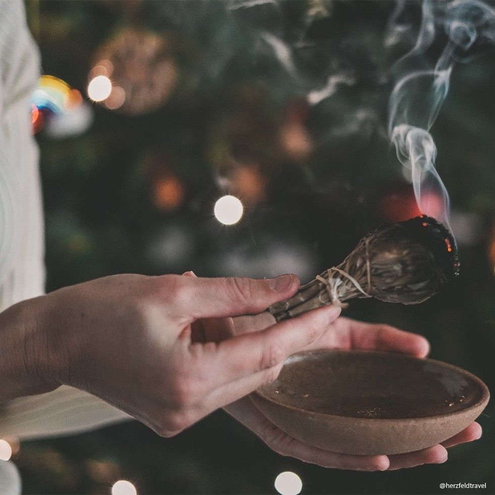 Hands holding a burning sage bundle over a bowl, with a Christmas tree in the background.