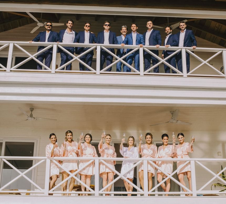 A happy bridal party on two balconies at Ibagari Hotel, groomsmen on top and bridesmaids below toasting with champagne