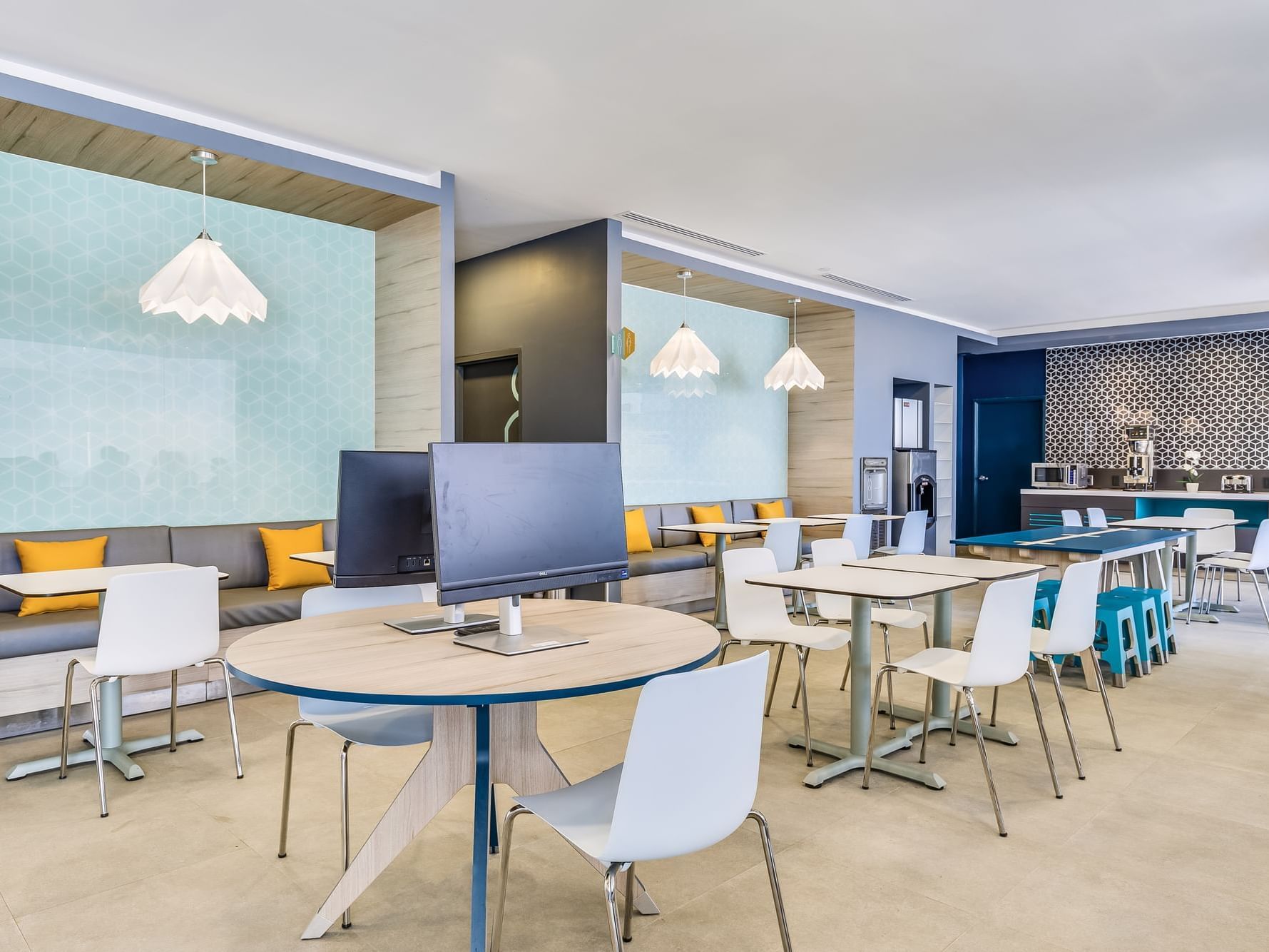 Modern office break room with tables, chairs, monitors, and a kitchen area at One Hotels