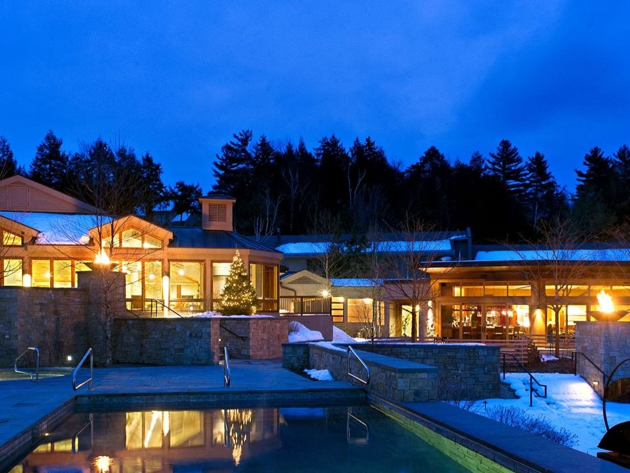 Nighttime view of a resort with a pool, lit buildings, and a festive Christmas tree, surrounded by snow and trees.