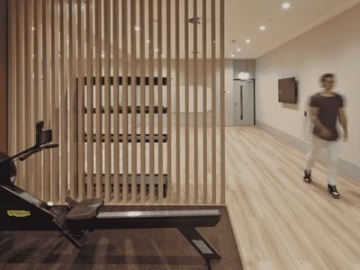 Person walking in a gym corridor with rowing machine, wall with vertical bars, and a TV on wall.