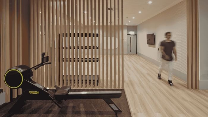 Modern gym room with rowing machine, wooden floor, and person walking in background.