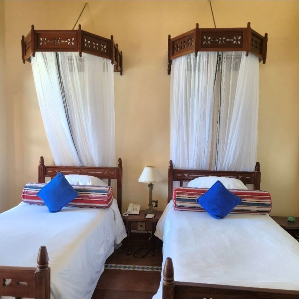 Two twin beds with blue pillows and mosquito nets in Standard Twin Room at Zanzibar Serena Hotel.