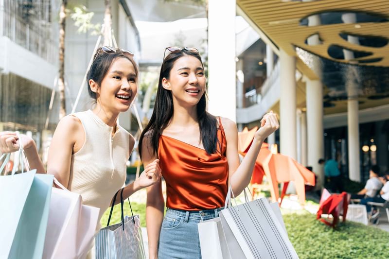 Two smiling Asian women with shopping bags walking outdoors at a modern shopping center near The Diplomat Resort