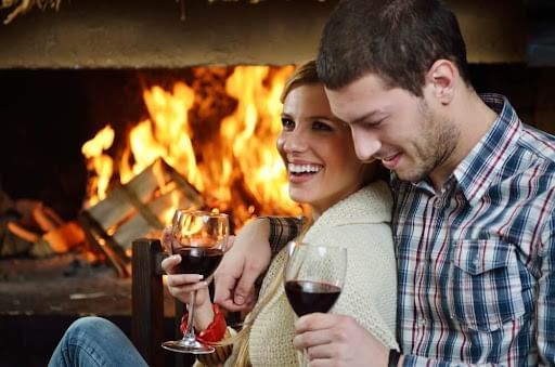 A couple happily cuddles by a roaring fireplace, each holding a glass of red wine at Cove Pocono Resorts