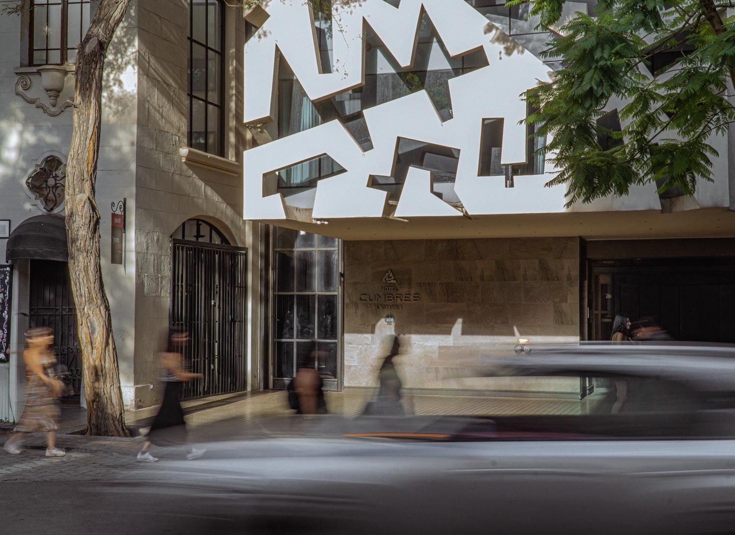 Fachada moderna del hotel con escultura blanca geométrica suspendida y peatones en movimiento en Cumbres Lastarria