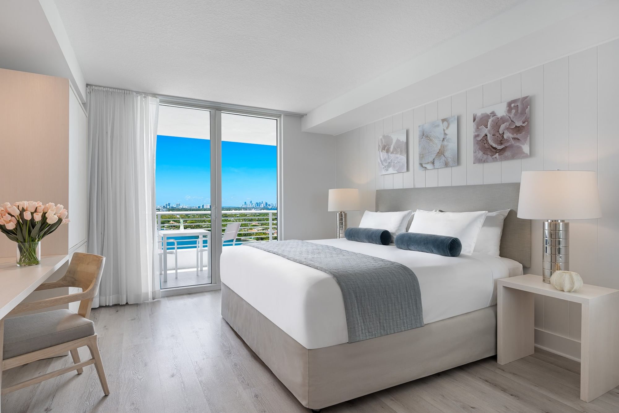 A wide view of a hotel room with a large bed, desk, and sliding glass door leading to a balcony with a city view.